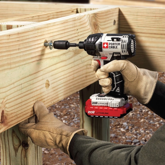 A person wearing work gloves uses a PORTER-CABLE 20V lithium cordless impact driver to tighten a screw into a wooden structure outdoors.