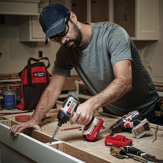 A person wearing a grey t-shirt and a blue cap is using a PORTER-CABLE cordless drill to assemble a wooden structure. Several PORTER-CABLE tools, batteries, and accessories are visible on the work surface, as well as a PORTER-CABLE branded tool bag in the background. The setting appears to be a workshop or a kitchen under construction.