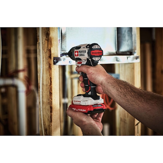A close-up of a person using a PORTER-CABLE brushless cordless impact driver to fasten a metal bracket to wooden framing. The tool features a 20V lithium battery. The scene appears to be part of a construction or renovation project.