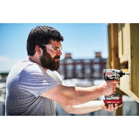 A person using a PORTER-CABLE 20V Lithium cordless impact driver to drill into a wooden surface outdoors. The person's face is blurred for privacy. The background shows a building and a clear sky.