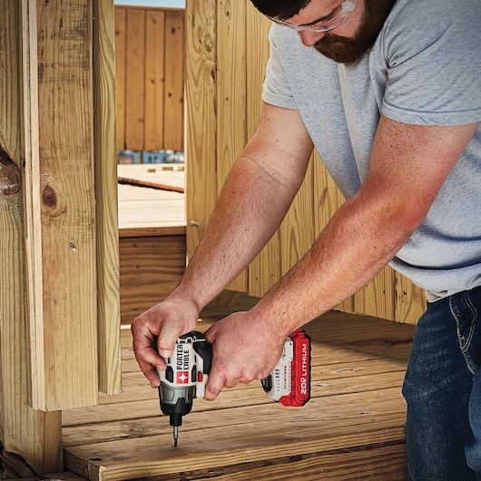 A person using a PORTER-CABLE 20V cordless impact driver to drill or fasten screws into wooden planks outdoors. The individual is wearing safety glasses and working on a construction project.