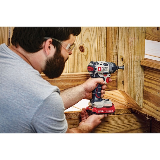 A person using a PORTER-CABLE 20V lithium cordless impact driver to drill into a wooden structure. The person's face is blurred for privacy.