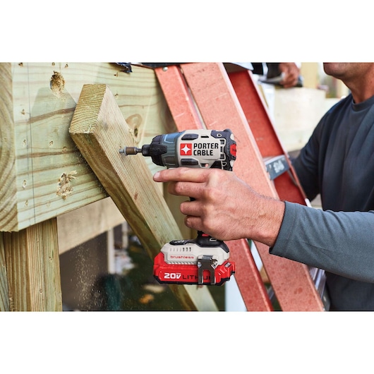A close-up of a person using a PORTER-CABLE 20V Lithium brushless cordless impact driver to fasten a screw into a piece of wood framing. There is a ladder and carpentry work in progress in the background.