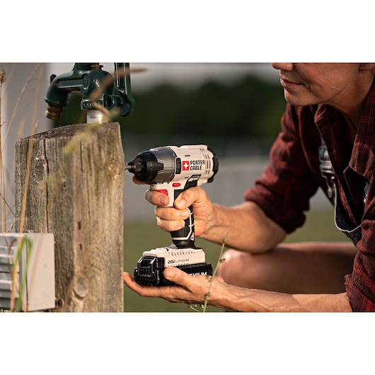 A person using a PORTER-CABLE 20V Lithium cordless impact driver to drive a screw into a wooden post outdoors. The tool is being held firmly, and gardening or construction equipment is visible nearby.