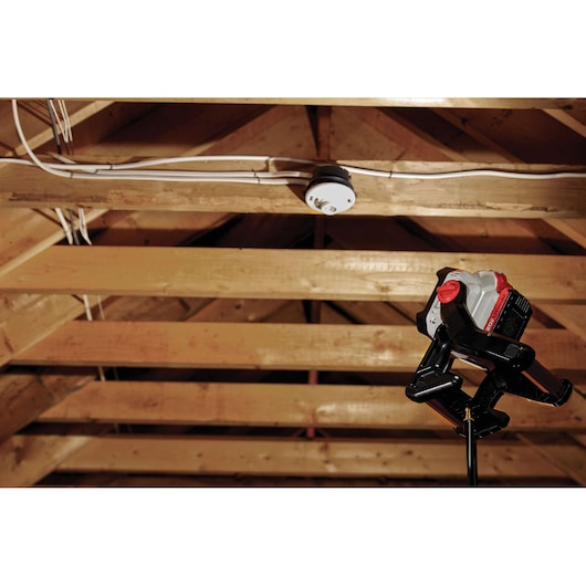 A PORTER-CABLE work light (model PCCL500B) is positioned on a stand, illuminating the ceiling structure of a wooden attic with visible beams and electrical wiring.