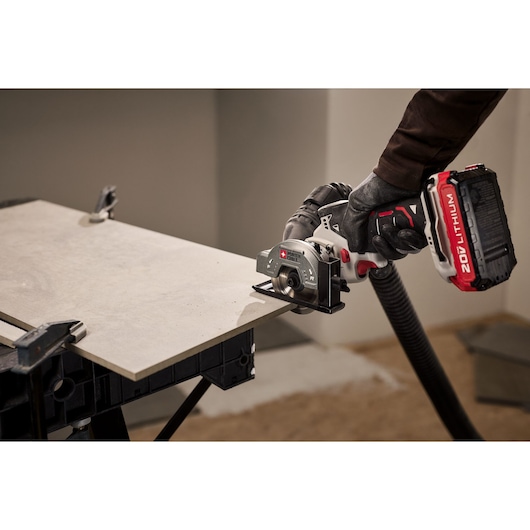 A person wearing gloves uses a PORTER-CABLE cordless tile saw with a 20V lithium battery to cut a ceramic tile placed on a workbench. The tile is secured with clamps and there is a vacuum hose attached to the saw for dust collection.