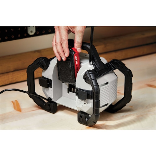 A PORTER-CABLE photography equipment device is shown with a person's hand inserting a 20V lithium battery. The device has a gray body with black protective handles and is placed on a wooden surface.