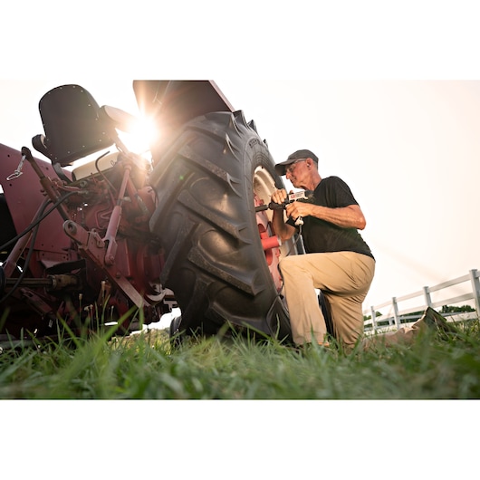 A person kneels beside a large tractor outdoors and uses a PORTER-CABLE power tool to work on the wheel, with sunlight shining in the background.