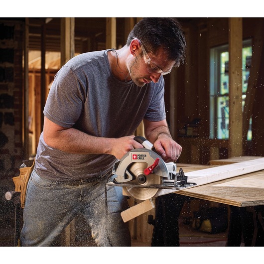 A person using a PORTER-CABLE circular saw to cut a wooden board inside a construction site. Sawdust is flying as the saw is in operation, and the operator is wearing a grey t-shirt and jeans.