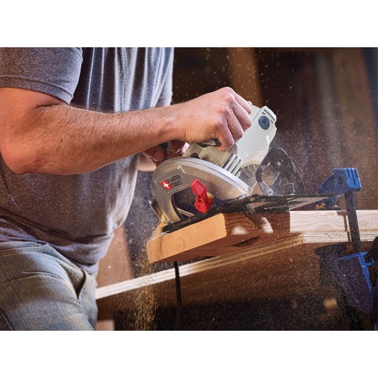 A person using a PORTER-CABLE circular saw (model PCE300) to cut a piece of wood, with sawdust flying during the operation. The wood is clamped in place for stability.
