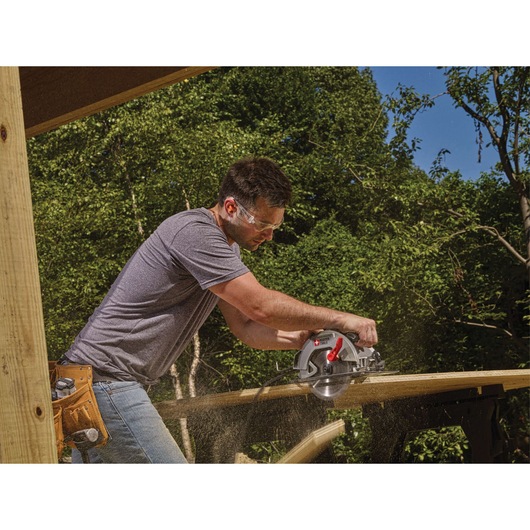 A person using a PORTER-CABLE circular saw to cut a wooden plank outdoors. The person is wearing a tool belt and sawdust is visible, with trees in the background.