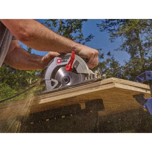 A PORTER-CABLE circular saw is being used to cut a sheet of plywood outdoors. Sawdust is visibly flying, and the person's hands are guiding the tool along the wood, with trees and a blue sky in the background.