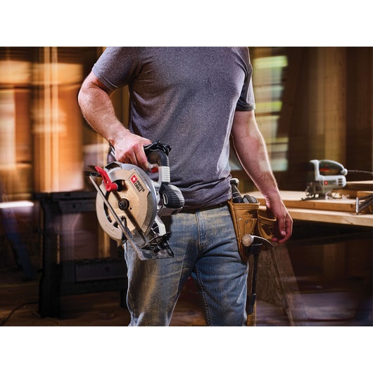 A person wearing a grey t-shirt and jeans is holding a PORTER-CABLE circular saw model PCE300 in a workshop setting. The person also has a tool belt with tools, and there is another power tool on a workbench in the background.