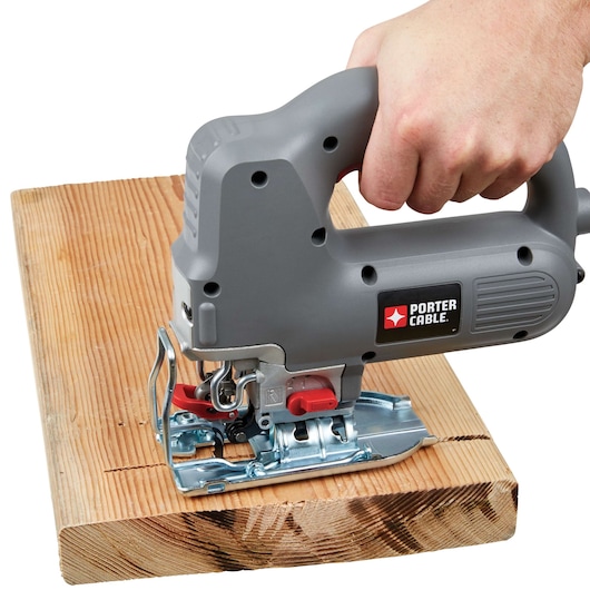 A PORTER-CABLE electric jigsaw is being used to cut a piece of wood. The tool is held by a person's hand and is creating a straight cut along the wood plank.