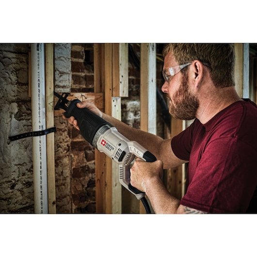 A person using a PORTER-CABLE reciprocating saw (model PCE360) to cut a PVC pipe in a construction or renovation setting. The person's face is blurred and the background shows exposed wooden framing and brick walls.