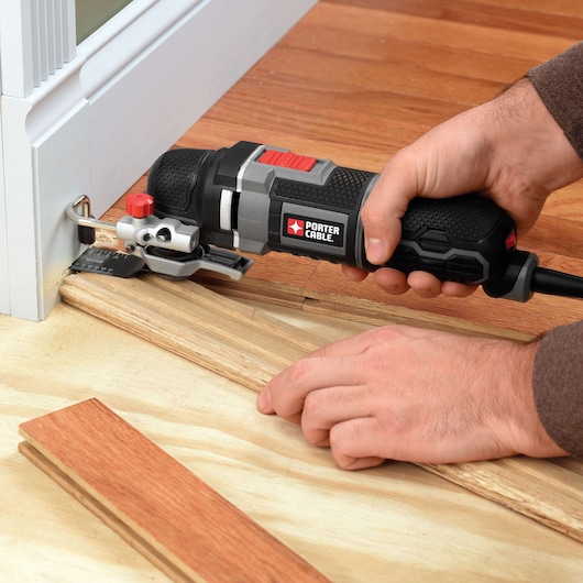A person using a PORTER-CABLE oscillating tool to cut wood flooring near a door frame. The tool is being used to trim the wood for a precise fit.