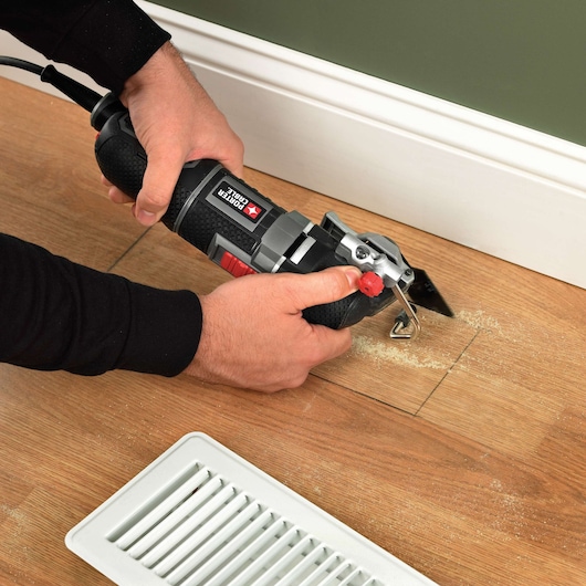 A person using a PORTER-CABLE oscillating tool to cut a rectangular section out of a wooden floor near a white floor vent and baseboard.