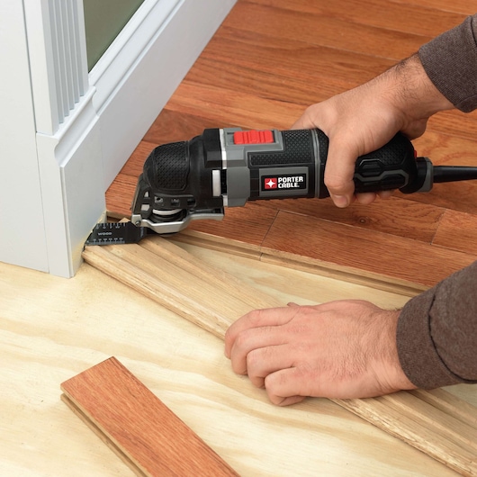 A person uses a PORTER-CABLE oscillating multi-tool to cut wood flooring near a door frame. The tool is positioned close to the base of the door trim, making precise cuts in the wood.