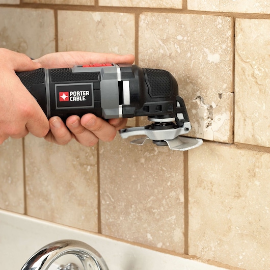 A person uses a PORTER-CABLE oscillating tool to cut or remove grout from beige ceramic tiles on a wall above a sink.
