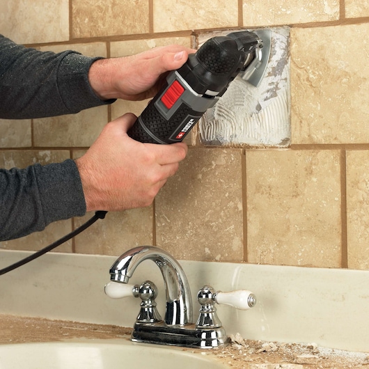 A person using a PORTER-CABLE oscillating power tool to remove tile adhesive from a wall above a bathroom sink with chrome faucet and beige tiles.