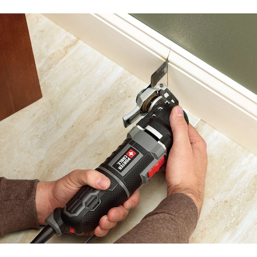 A PORTER-CABLE oscillating multi-tool is being used to cut a baseboard. The tool is held in both hands and positioned against the white baseboard, with a cutting blade making a precise incision. The surface is a tiled floor.