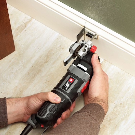 A person using a PORTER-CABLE PCE605K oscillating tool to cut into a white baseboard in a room with light-colored flooring.