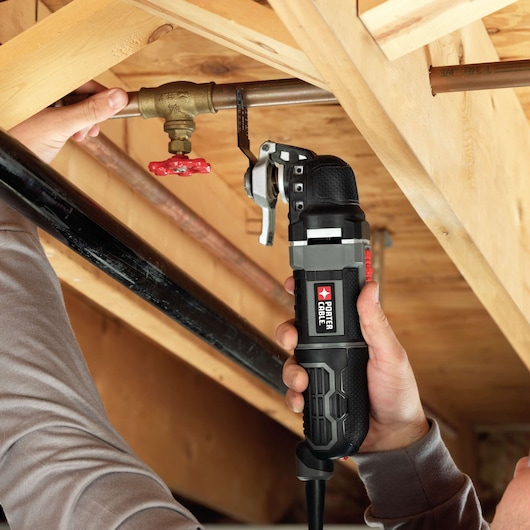 A PORTER-CABLE oscillating multi-tool being used to cut a metal pipe near a valve in a wooden ceiling framework.