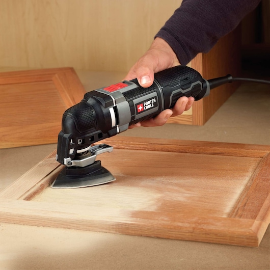 A person is holding a PORTER-CABLE electric multi-tool, sanding the surface of a wooden frame. The tool is plugged in and in use on a workbench.