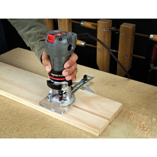 A handheld PORTER-CABLE router being used to cut grooves into a piece of wood on a workbench. The tool is held by a person and is plugged in for operation. Various woodworking tools are visible in the background.