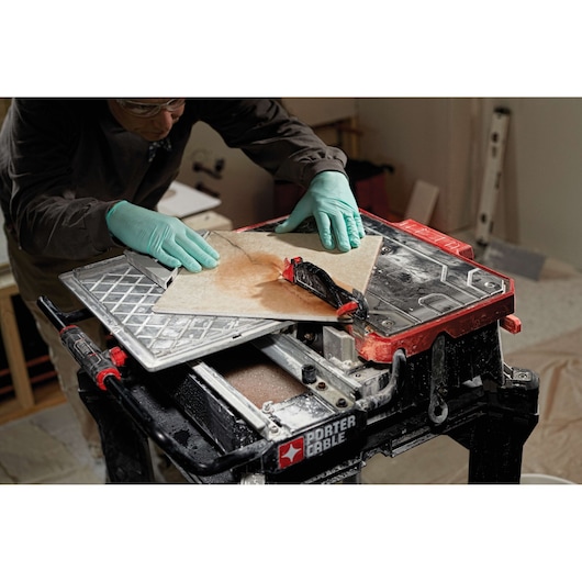 A person wearing green gloves is using a PORTER-CABLE PCE980 tile saw to cut a large ceramic tile in a workshop or construction environment. The saw is mounted on a stand, and there are tools and equipment visible in the background.