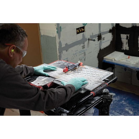 A person wearing green gloves uses a PORTER-CABLE PCE980 tile saw to cut a ceramic tile in a workshop setting. The saw is placed on a stand, and construction materials are visible in the background.