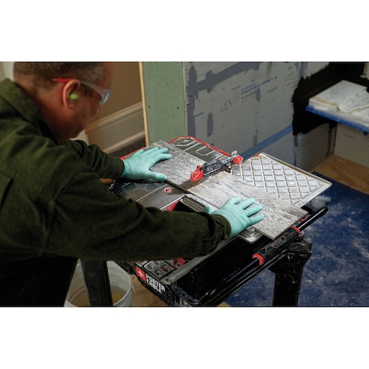 A person wearing green gloves uses a PORTER-CABLE tile saw (model PCE980) to cut a piece of tile on a worktable. The saw is set up indoors, in front of a wall under construction.