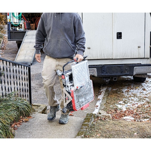 A person wearing a gray hoodie and khaki pants is carrying a PORTER-CABLE PCE980 tool, walking up a sidewalk. The tool appears to be a tabletop saw or tile cutter. There is a truck in the background with equipment and supplies visible.