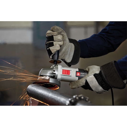 A person wearing protective gloves is using a PORTER-CABLE angle grinder to cut or grind a metal pipe. Sparks are visible as the tool operates, indicating active metalwork.