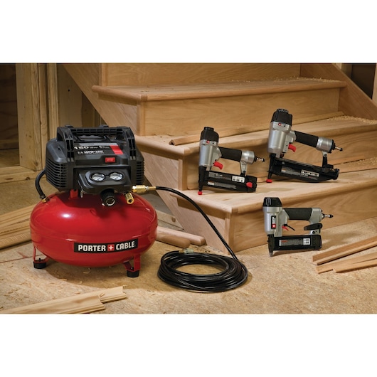 PORTER-CABLE PCFP3KIT air compressor kit displayed with three pneumatic tools and a coiled air hose, set in a woodworking environment with unfinished wooden stairs and various wood pieces.