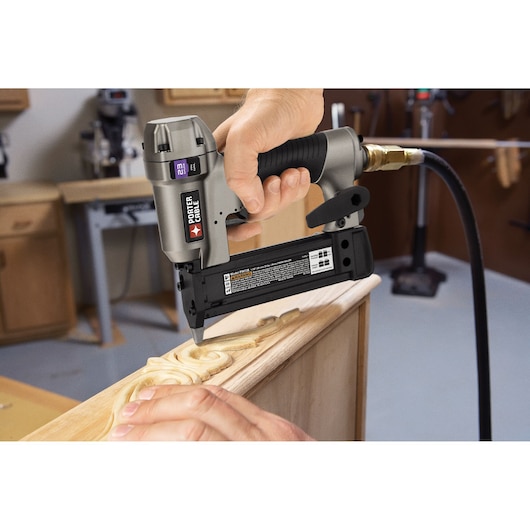 A close-up of a PORTER-CABLE pneumatic pin nailer being used to fasten decorative wood trim onto a piece of furniture in a woodworking shop.