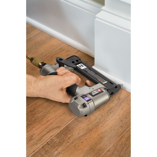 A PORTER-CABLE PIN138 pneumatic pin nailer being used to install white baseboard trim on a wooden floor.