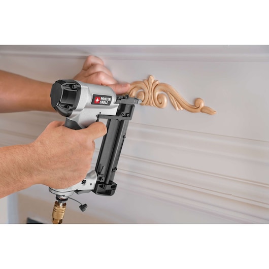 A person uses a PORTER-CABLE branded pneumatic pin nailer to attach decorative molding to a white wall.
