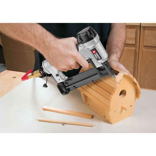 A person is using a PORTER-CABLE pneumatic pin nailer to assemble a wooden birdhouse on a work surface. Two wooden strips and a small screw are laying nearby.