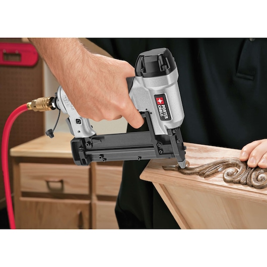 A close-up of a PORTER-CABLE PIN138 pneumatic pin nailer being used to attach decorative wood trim to a wooden furniture piece. The tool is connected to a red air hose, and a person's hand is holding it firmly against the wood.