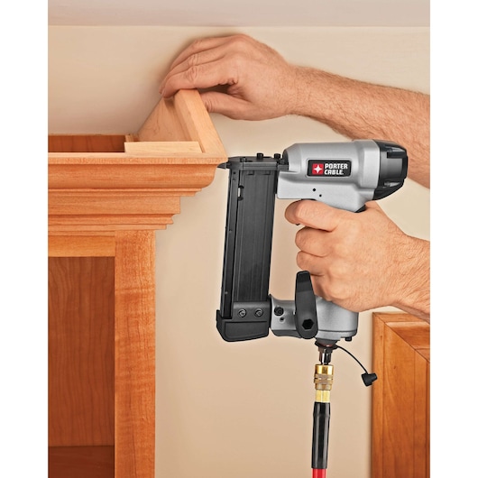 A close-up of a person using a PORTER-CABLE pneumatic pin nailer (model PIN138) to attach wood trim or molding to a cabinet.