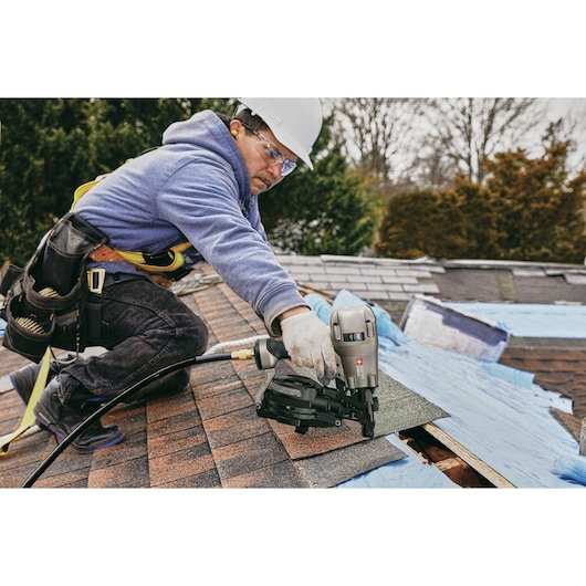 A person wearing gloves, a safety harness, and a hard hat kneels on a roof while using a PORTER-CABLE RN175C pneumatic nailer to install shingles. Roofing materials and blue underlayment are visible on the roof.
