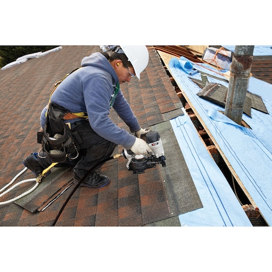 A construction worker wearing safety gear installs roof shingles using a PORTER-CABLE pneumatic nail gun on a rooftop. The worker appears to be kneeling on the roof and is surrounded by roofing materials.