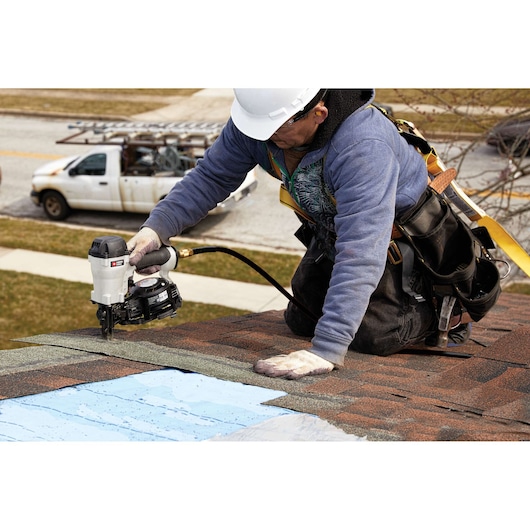 A person wearing a safety harness and hard hat uses a PORTER-CABLE pneumatic nailer (SKU: RN175C) to install roofing shingles on a house roof. Construction tools and a white pickup truck with a ladder are visible in the background.