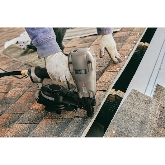 A person wearing gloves is using a PORTER-CABLE pneumatic roofing nailer (model RN175C) to install shingles on a roof.