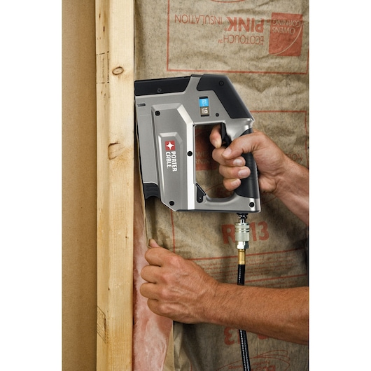 A PORTER-CABLE pneumatic stapler being used to secure insulation material to a wooden frame in a construction setting. The tool is held by a hand and connected to an air hose.