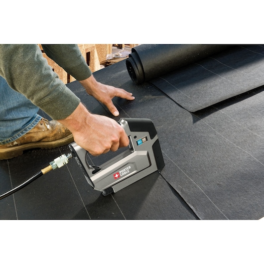 A person using a PORTER-CABLE pneumatic stapler (model TS056) to secure roofing felt onto a roof surface. The stapler is connected to an air hose and the person is wearing work boots.